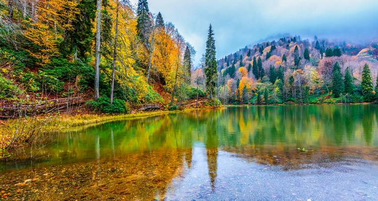 Calm mountain lake reflecting colorful autumn trees and misty hills.