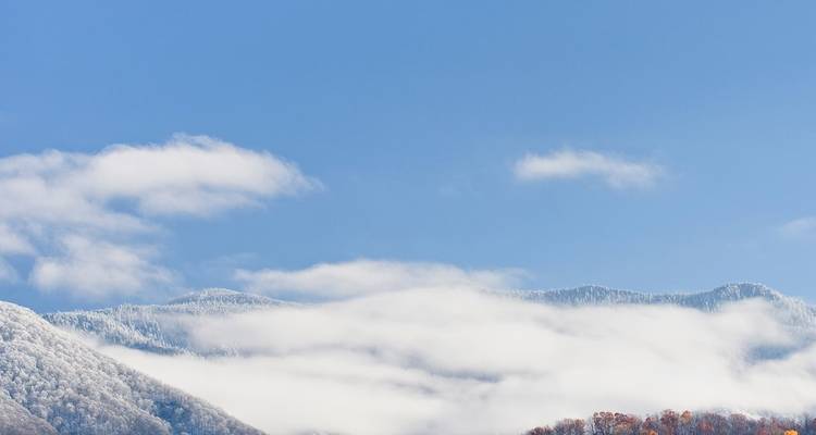 Snow-capped ridges of the Great Smoky Mountains peek through rolling clouds beneath a bright blue sky