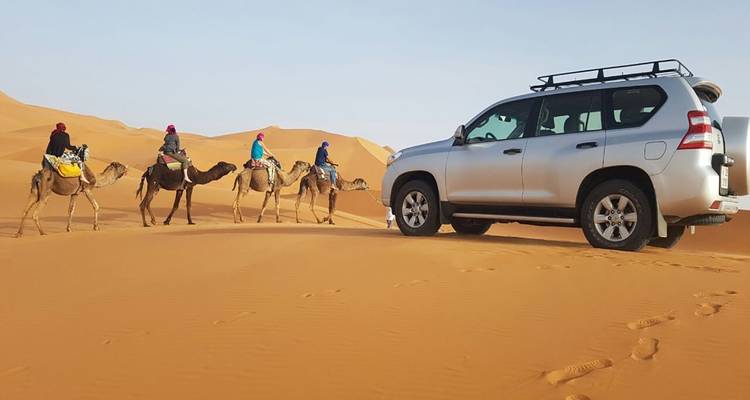 Gruppe von Menschen, die auf Kamelen in einer Wüste reiten, mit einem Geländewagen in der Nähe.