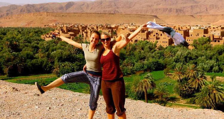 Zwei Frauen posieren fröhlich mit einer malerischen Stadt und Grün im Hintergrund.