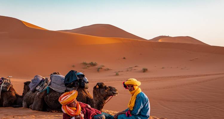 Zwei Menschen in bunter Kleidung sitzen mit Kamelen in einer weiten Wüstenlandschaft.