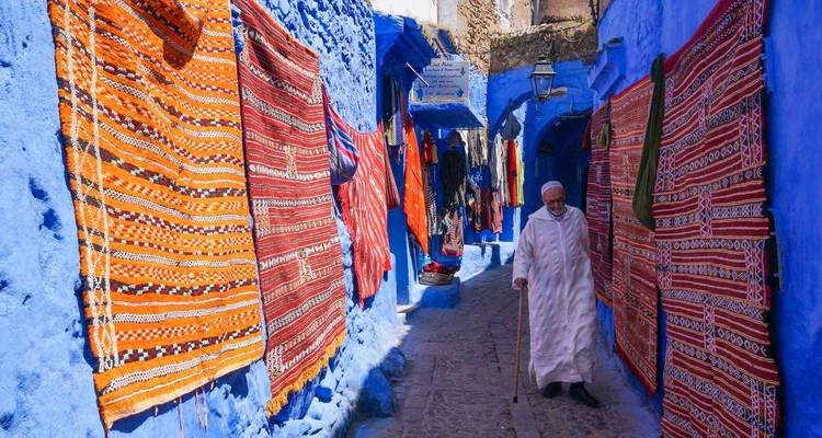 Man die door een levendige straat in Chefchaouen loopt met kleurrijke tapijten.