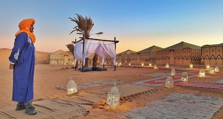 Man in traditional clothing in a desert camp with candles and palm trees at sunset.