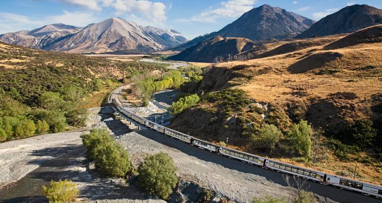 Long passenger train snakes through a mountain valley under clear blue skies in New Zealand.