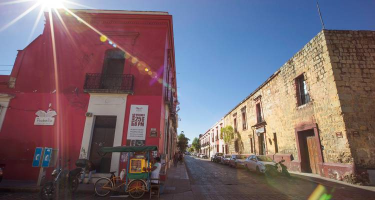 El sol de la mañana brilla sobre una calle colonial llena de color, rodeada de edificios vibrantes y adoquines en Oaxaca.