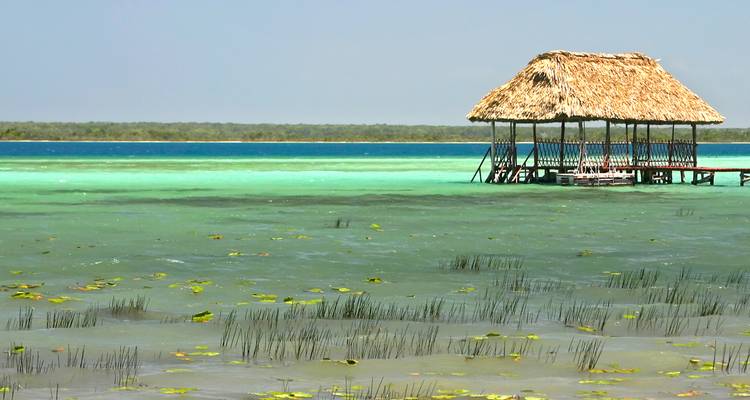 Strohdachte Palapa-Hütte auf Stelzen über den türkisfarbenen Gewässern und der seerosengefleckten Lagune von Bacalar.