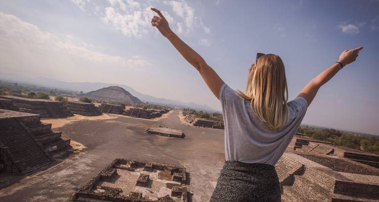 Una joven con los brazos levantados contempla las antiguas pirámides y plazas de un gran sitio arqueológico bajo un cielo despejado.
