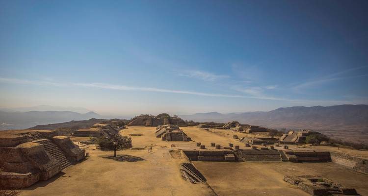 Vista amplia de una antigua ciudad zapoteca en la cima de una colina con terrazas, pirámides y montañas circundantes bajo un cielo azul.