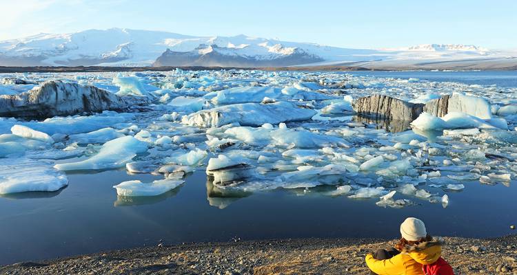 Voyageur vêtu d'une veste jaune assis près d'une lagune glacée couverte de blocs de glace bleue et de montagnes enneigées au loin.