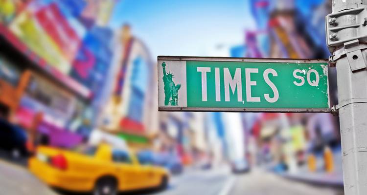 Green Times Square street sign with blurred yellow taxi and colorful billboards in the background.