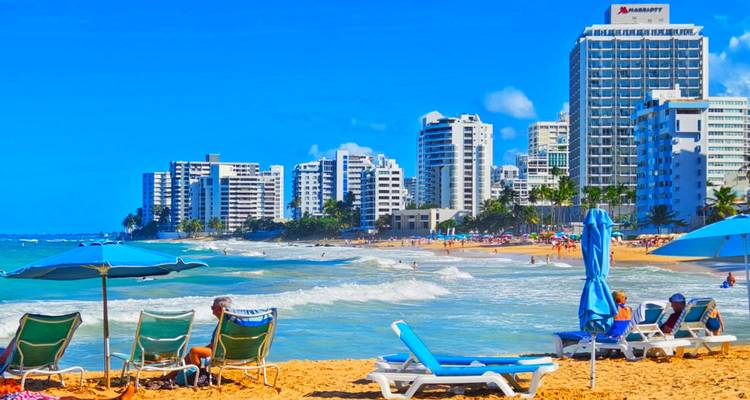 Playa concurrida con bañistas tomando el sol y edificios de gran altura.