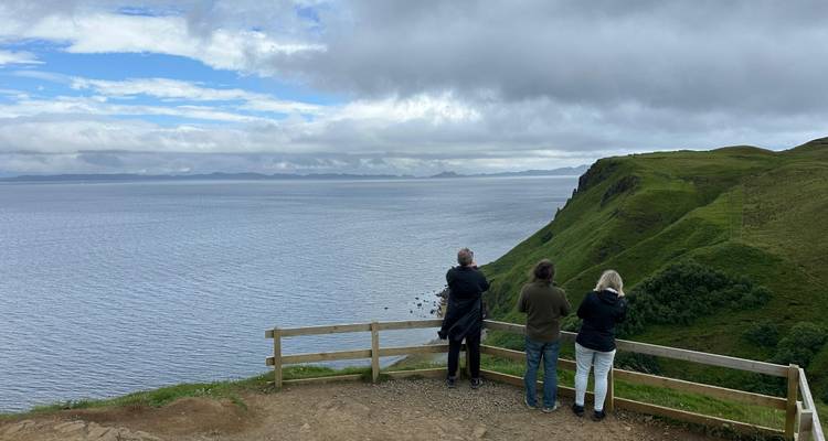 Trois voyageurs à la limite d'une falaise, appuyés contre une barrière en bois, contemplant la vaste mer et les îles lointaines sous un ciel dramatique.