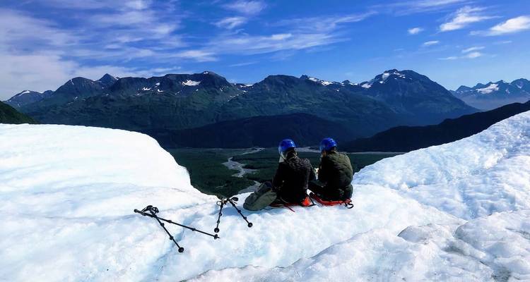 Two people sitting on a glacier edge with trekking poles, taking in the landscape.