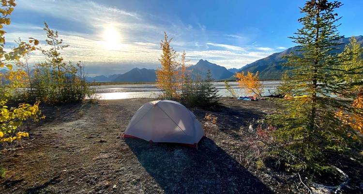 Tent pitched on a lakeside with autumn foliage and mountains.