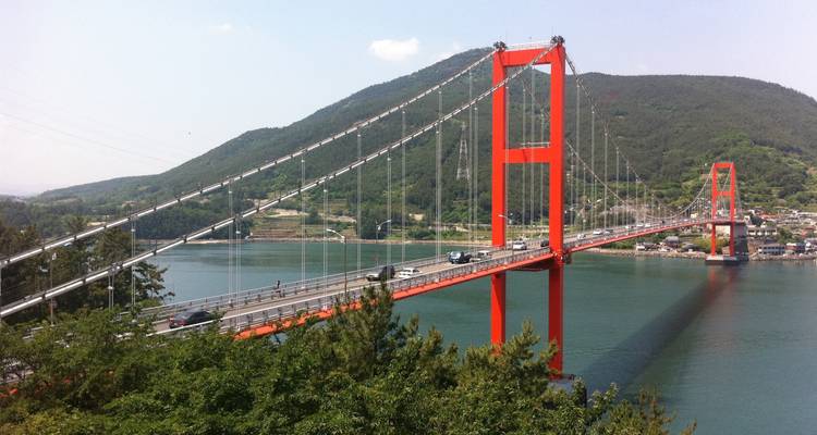 Felrode hangbrug over turquoise water met beboste bergen erbuiten op een zonnige dag.
