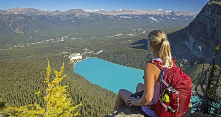 Wanderer sitzt auf einer Klippe mit Blick auf einen türkisfarbenen See, umgeben von Bergen.