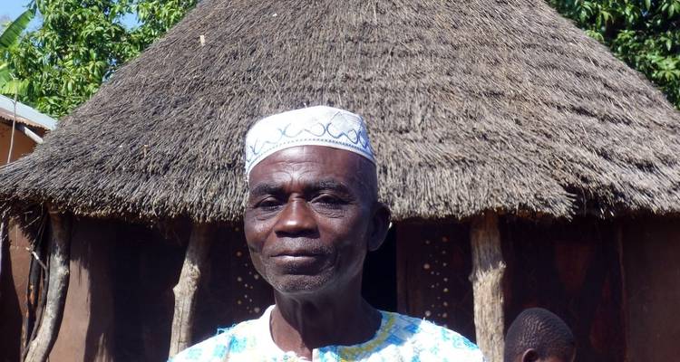 Man standing in front of a traditional thatched-roof hut.