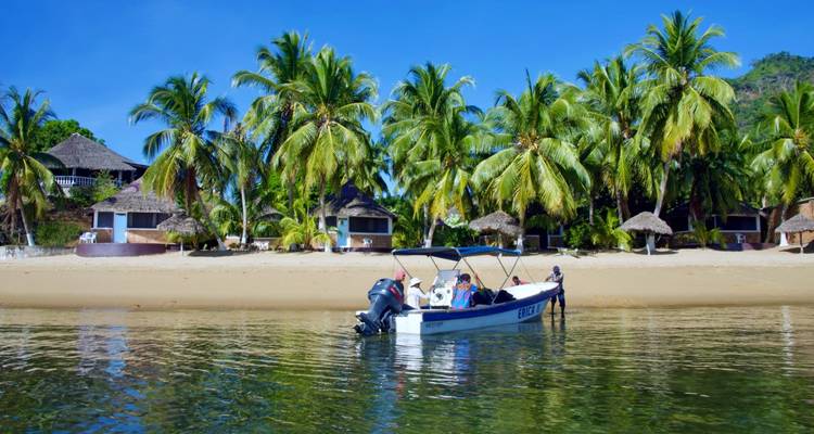 A boat on a tropical beach with palm trees and straw huts in the background.