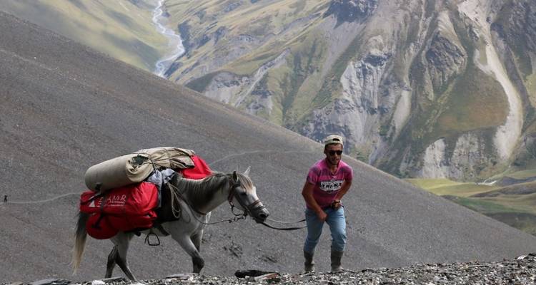 Homme menant un cheval chargé d'équipement sur un sentier accidenté.