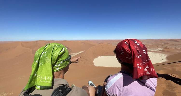 Two travelers in headscarves admire sweeping red sand dunes under a deep blue desert sky.