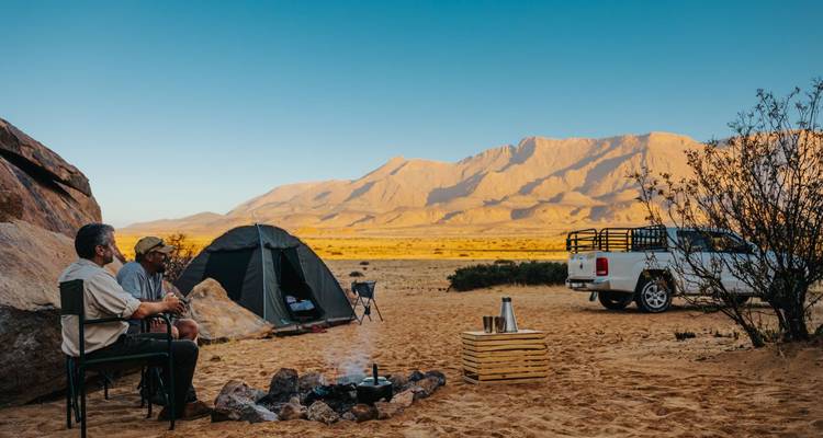 Desert campsite with tent, pickup truck and campfire against golden mountain backdrop at dawn.