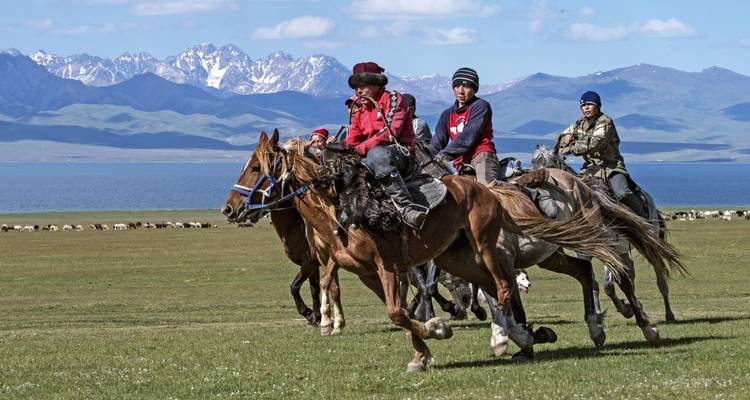 Group of people riding horses across a grassy plain near mountains.