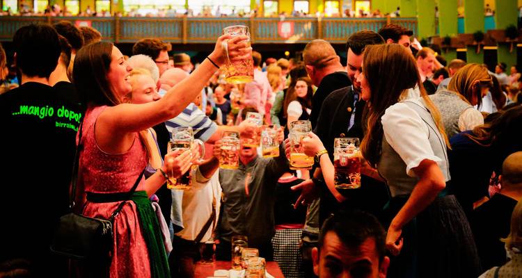 Joyful crowd toasting large beer steins inside a festive hall, likely during Oktoberfest.