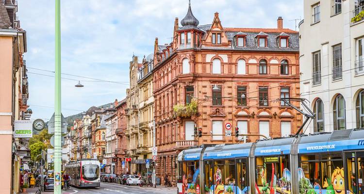 Trams pass colorful 19th-century buildings on a busy shopping street lined with overhead cables.