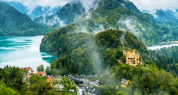 Misty aerial view of emerald lake and yellow Hohenschwangau Castle nestled in dense forested hills.