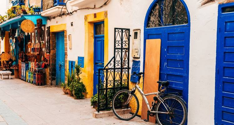 Rue blanchie à la chaux à Essaouira avec des portes bleu vif, une bicyclette et un étal de marché coloré.