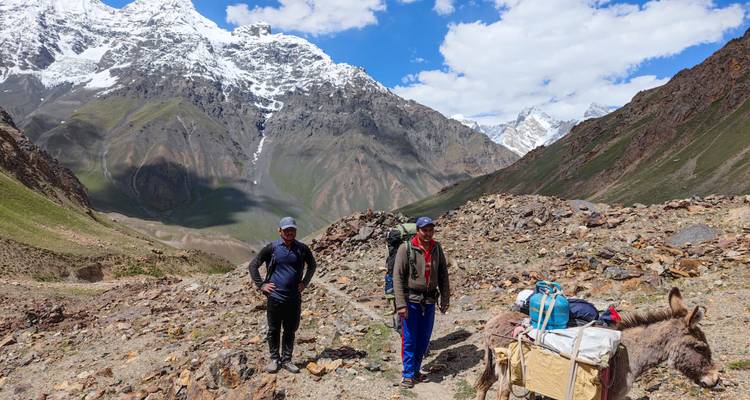 Two trekkers with pack donkey standing on rocky alpine trail amid high peaks.