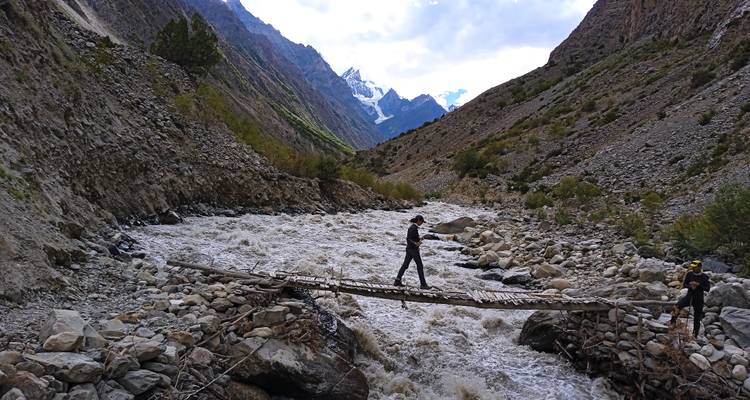 Adventurers crossing a narrow wooden bridge over a fast-flowing mountain river with snowy peaks ahead