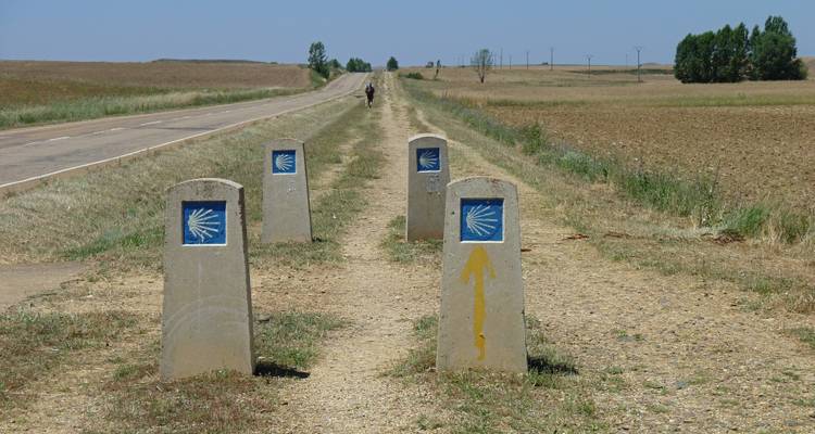 Un sentier de terre avec des marqueurs arborant le symbole du Camino de Santiago, et une silhouette au loin.