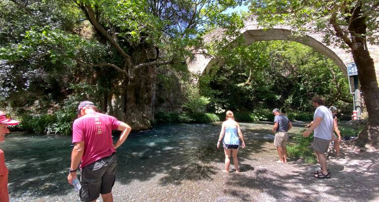 Des gens qui pataugent dans une rivière peu profonde sous un pont de pierre entouré d'une végétation luxuriante.