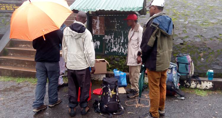 Gruppe, die bei einem Gebäude mit Taschen und Regenschirmen in regnerischer Umgebung steht.