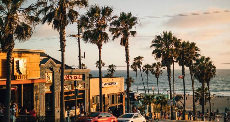 Vibrant beachfront street scene with palm trees, shops and diners overlooking the ocean.