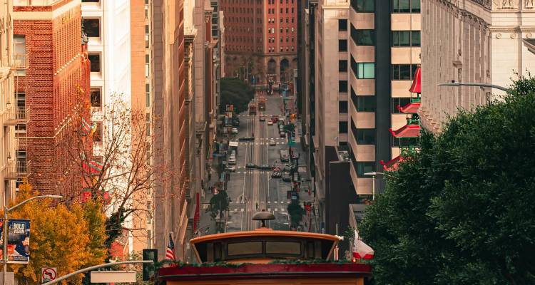 A steep downtown street in San Francisco with a historic red cable car heading downhill between tall buildings in warm light.