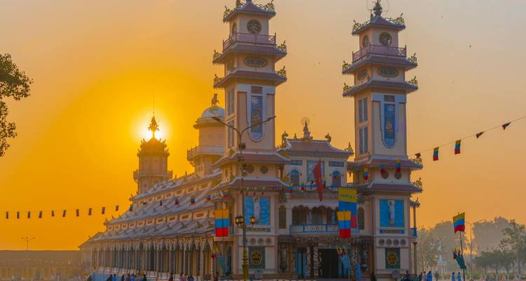 Le Temple Cao Dai brille devant un coucher de soleil doré, ses tours ornées encadrées par des drapeaux flottants.