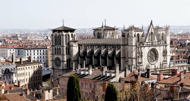 Steinerne gotische Kathedrale von Lyon, die über den Terrakotta-Dächern der Altstadt unter hellem Himmel thront.