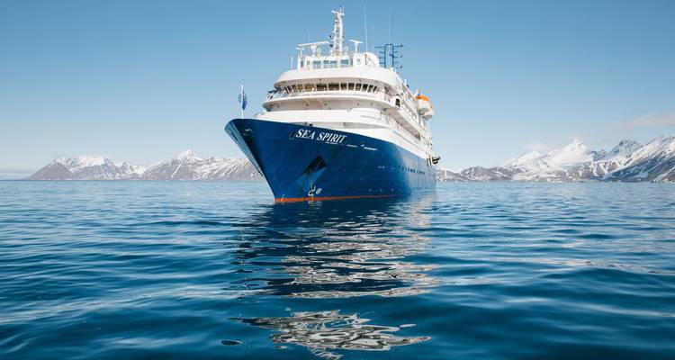 El barco expedicionario 'Sea Spirit' está navegando en las tranquilas aguas azules del Ártico con montañas cubiertas de nieve a lo lejos.