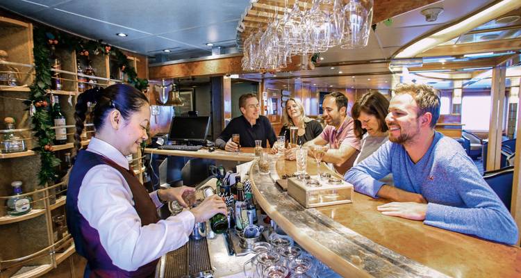 Los viajeros disfrutan de bebidas en un acogedor bar del barco mientras un cantinero prepara cócteles.