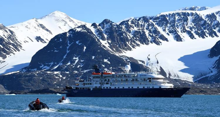 Dos pequeños Zodiacs se alejan a toda velocidad de un crucero de expedición anclado con un dramático telón de fondo de montañas cubiertas de nieve.