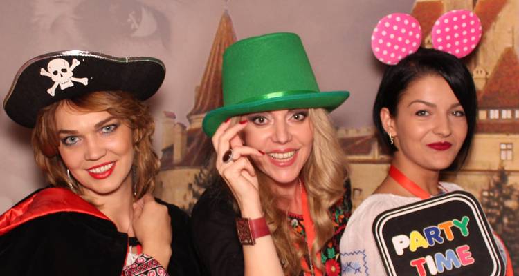 Three women in colourful Halloween costumes posing cheerfully in a photo-booth with a medieval castle backdrop.