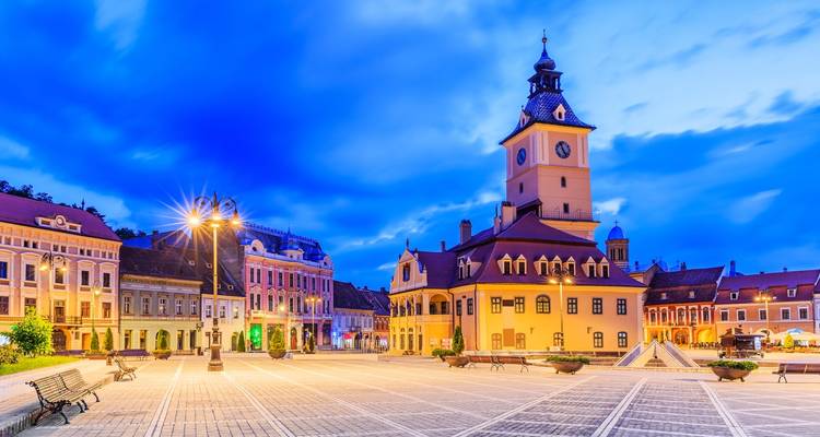 Evening panorama of Brasov’s Council Square with the illuminated Council House and colourful baroque facades under a vivid sky.