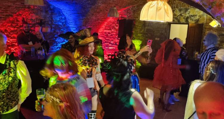 Costumed guests dance and mingle in a vaulted stone cellar lit by colourful party lights.