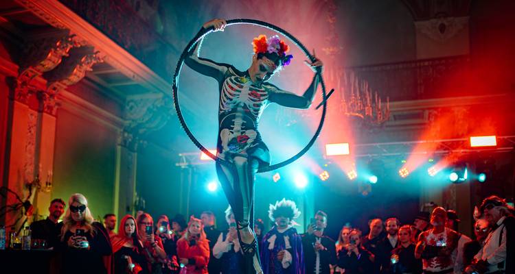 Costumed performer in skeleton bodysuit spins inside a hoop on stage at a vibrant themed party.