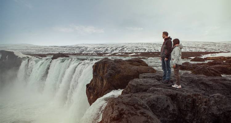 Una pareja se para en una saliente rocosa mirando hacia una amplia cascada brumosa en un paisaje invernal.
