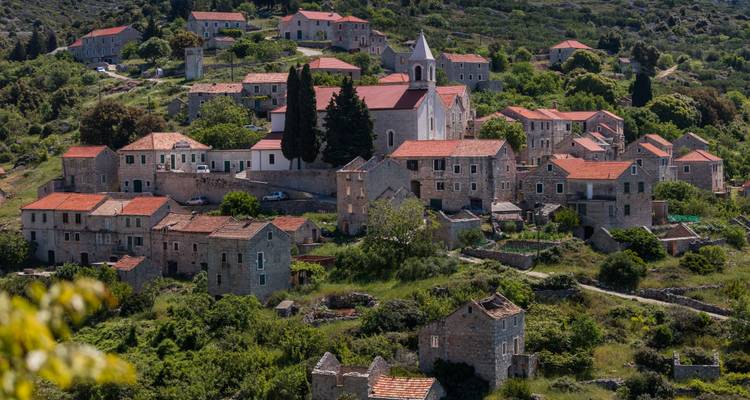 A picturesque stone village with red-tiled roofs and a white hilltop church surrounded by lush green hills on a Croatian island.