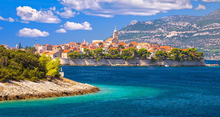 Korčula’s fortified old town with orange roofs sits on a rocky peninsula above turquoise Adriatic waters, backed by rugged mountains and scattered clouds.