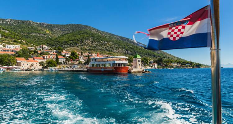 Croatian flag flying from a boat leaving a coastal village behind.
